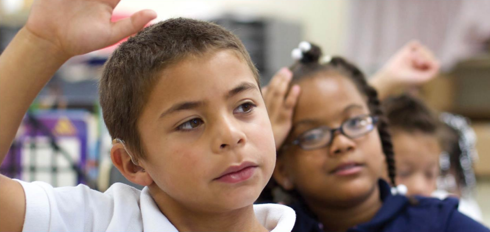 Image of student holding up his hand in a classroom while another student looks confused. 